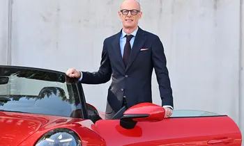 Ein glatzköpfiger Mann mit dunkler Brille, dunkelblauem Anzug und hellblauem Hemd steht lächelnd neben einem roten Sportwagen mit geöffneter Tür.
