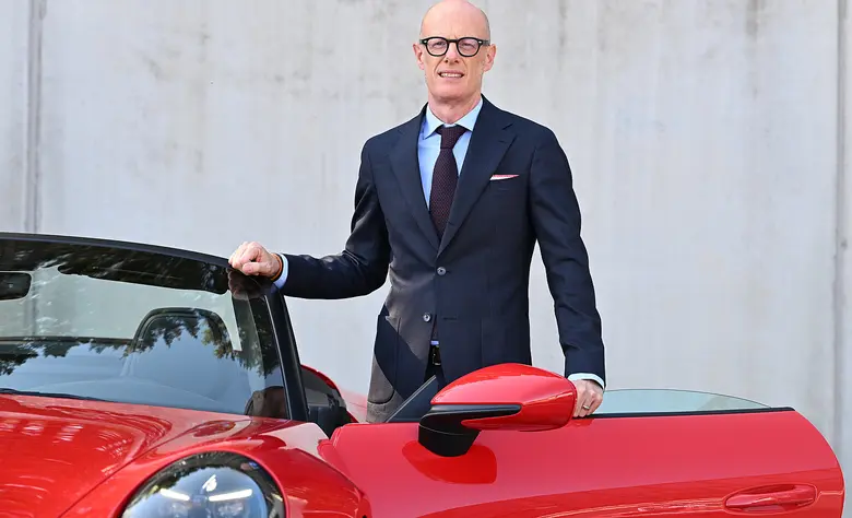 Ein glatzköpfiger Mann mit dunkler Brille, dunkelblauem Anzug und hellblauem Hemd steht lächelnd neben einem roten Sportwagen mit geöffneter Tür.