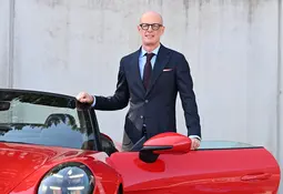 Ein glatzköpfiger Mann mit dunkler Brille, dunkelblauem Anzug und hellblauem Hemd steht lächelnd neben einem roten Sportwagen mit geöffneter Tür.