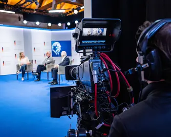 Fernsehkamera vor einer Bühne mit drei Personen bei einer Paneldiskussion.
