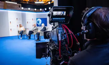Fernsehkamera vor einer Bühne mit drei Personen bei einer Paneldiskussion.