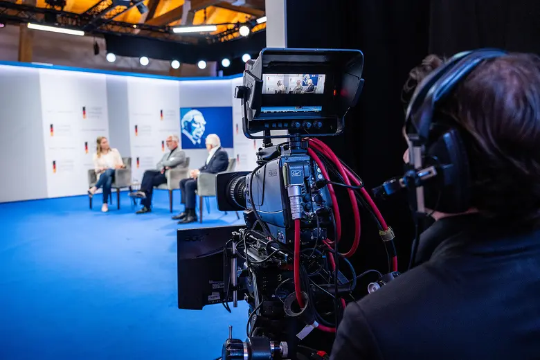 Fernsehkamera vor einer Bühne mit drei Personen bei einer Paneldiskussion.