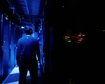 Eine Person von hinten in einem dunklen Serverraum, beleuchtet von blauem Licht der Serverracks.