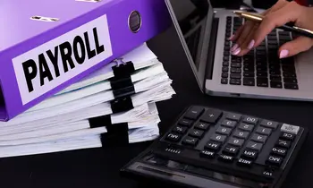 Lilafarbener Ordner mit der Aufschrift Payroll auf einem Stapel Dokumente, neben einem Taschenrechner und einem Laptop.