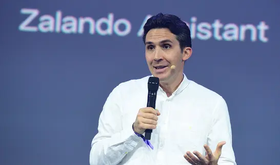 Ein Mann mit dunklen Haaren im weißen Hemd spricht mit einem Mikrofon in der Hand vor einem blauen Bildschirm, auf dem der Schriftzug Zalando Assistant zu sehen ist.