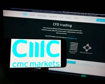 Eine Hand hält ein Smartphone mit dem CMC Markets Logo auf dem Display, im Hintergrund ist ein Monitor mit der CMC-Handelsplattform zu sehen.