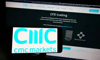 Eine Hand hält ein Smartphone mit dem CMC Markets Logo auf dem Display, im Hintergrund ist ein Monitor mit der CMC-Handelsplattform zu sehen.
