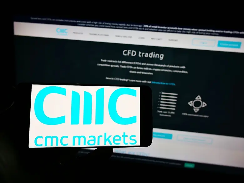 Eine Hand hält ein Smartphone mit dem CMC Markets Logo auf dem Display, im Hintergrund ist ein Monitor mit der CMC-Handelsplattform zu sehen.