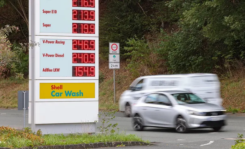Digitale Preistafel einer Shell-Tankstelle mit Kraftstoffpreisen, davor fährt ein silberfarbenes Auto mit Bewegungsunschärfe vorbei, im Hintergrund Bäume und ein Verkehrsschild.