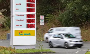 Digitale Preistafel einer Shell-Tankstelle mit Kraftstoffpreisen, davor fährt ein silberfarbenes Auto mit Bewegungsunschärfe vorbei, im Hintergrund Bäume und ein Verkehrsschild.