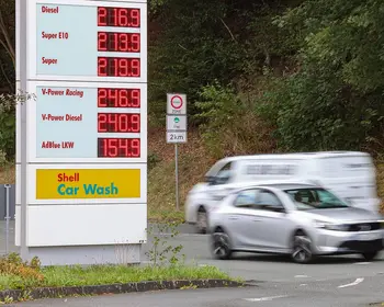 Digitale Preistafel einer Shell-Tankstelle mit Kraftstoffpreisen, davor fährt ein silberfarbenes Auto mit Bewegungsunschärfe vorbei, im Hintergrund Bäume und ein Verkehrsschild.