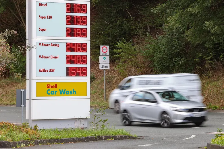 Digitale Preistafel einer Shell-Tankstelle mit Kraftstoffpreisen, davor fährt ein silberfarbenes Auto mit Bewegungsunschärfe vorbei, im Hintergrund Bäume und ein Verkehrsschild.