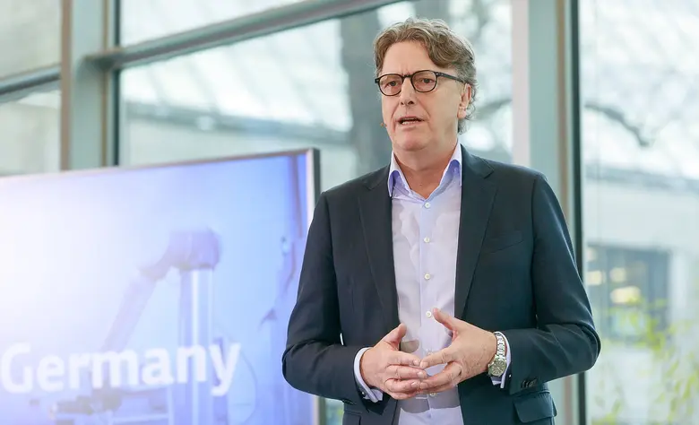 Mann im Anzug mit Brille und Headset vor einem Bildschirm.