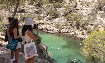 Zwei Frauen von hinten fotografieren mit dem Smartphone eine türkisfarbene Bucht von einem Felsweg aus.