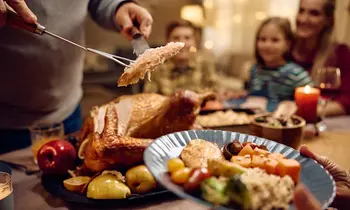Eine Person schneidet einen Braten am Esstisch an, im Hintergrund sitzen weitere Menschen.