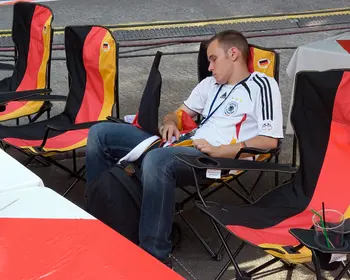 Mann in weißem DFB-Trikot schläft auf einem Campingstuhl, umgeben von leeren schwarz-rot-goldenen Klappstühlen auf einer Fanmeile.