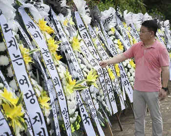 Ein Mann im rosafarbenen Poloshirt beugt sich zu einem von vielen aufgereihten weißen Trauerkränzen mit koreanischer Beschriftung und ordnet die gelben und weißen Blüten daran.