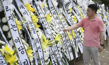 Ein Mann im rosafarbenen Poloshirt beugt sich zu einem von vielen aufgereihten weißen Trauerkränzen mit koreanischer Beschriftung und ordnet die gelben und weißen Blüten daran.