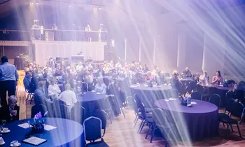 Zahlreiche Gäste sitzen an runden Tischen mit dunklen Tischdecken in einer großen Eventhalle, durch die breite Lichtstrahlen von der Bühne fallen.