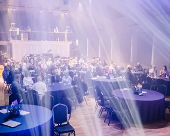 Zahlreiche Gäste sitzen an runden Tischen mit dunklen Tischdecken in einer großen Eventhalle, durch die breite Lichtstrahlen von der Bühne fallen.