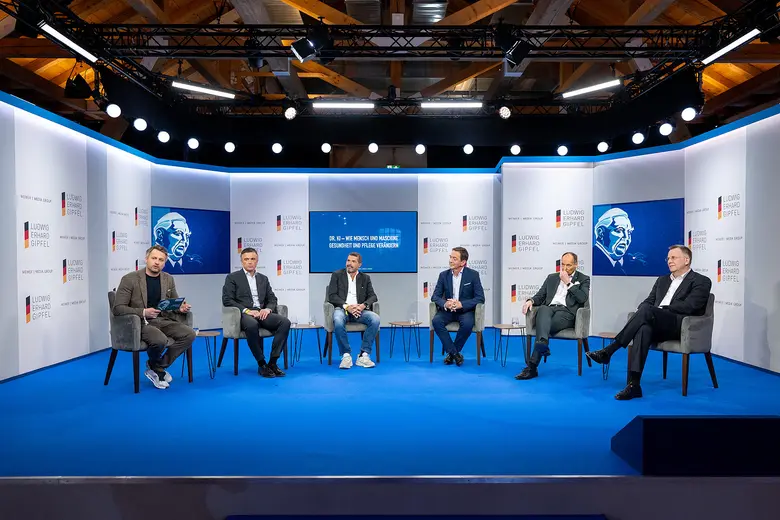 Sechs Männer sitzen bei einer Podiumsdiskussion auf einer blau beleuchteten Bühne.