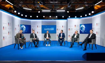 Sechs Männer sitzen bei einer Podiumsdiskussion auf einer blau beleuchteten Bühne.