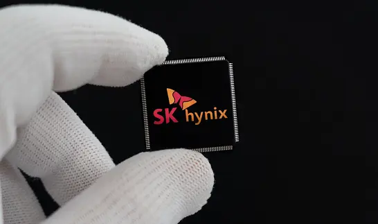 Zwei Finger in weißen Handschuhen halten einen schwarzen Chip mit dem Logo von SK hynix vor schwarzem Hintergrund.