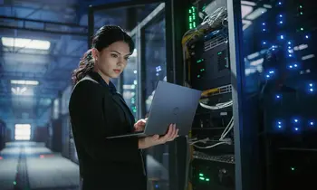 Eine Frau in dunkler Kleidung steht seitlich vor einem Serverschrank in einem Rechenzentrum und blickt konzentriert auf einen aufgeklappten Laptop in ihren Händen.