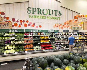 Blick in ein Ladenregal mit Obst und Gemüse unter dem Schriftzug Sprouts Farmers Market, im Hintergrund ein Kunde beim Einkaufen.