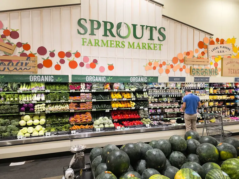 Blick in ein Ladenregal mit Obst und Gemüse unter dem Schriftzug Sprouts Farmers Market, im Hintergrund ein Kunde beim Einkaufen.