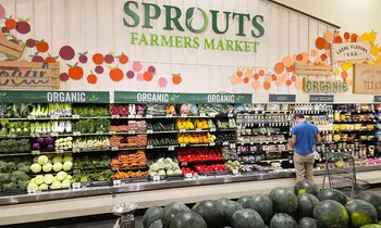 Blick in ein Ladenregal mit Obst und Gemüse unter dem Schriftzug Sprouts Farmers Market, im Hintergrund ein Kunde beim Einkaufen.