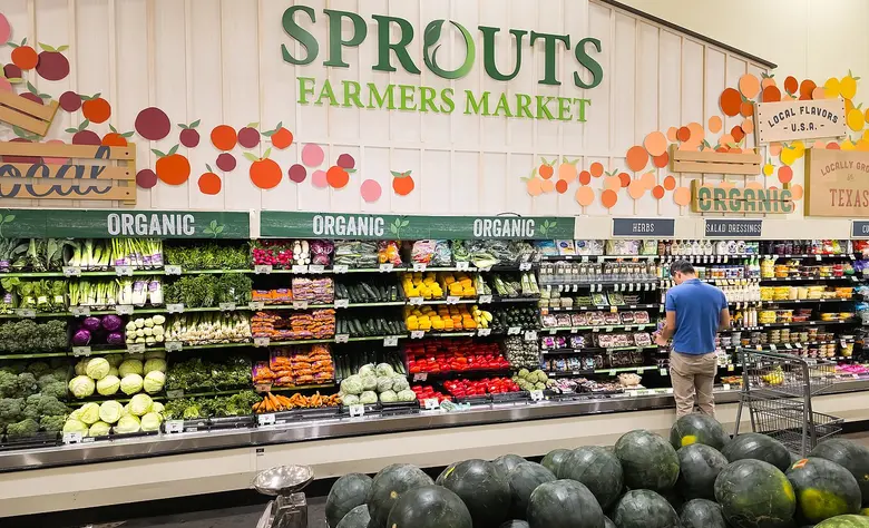 Blick in ein Ladenregal mit Obst und Gemüse unter dem Schriftzug Sprouts Farmers Market, im Hintergrund ein Kunde beim Einkaufen.