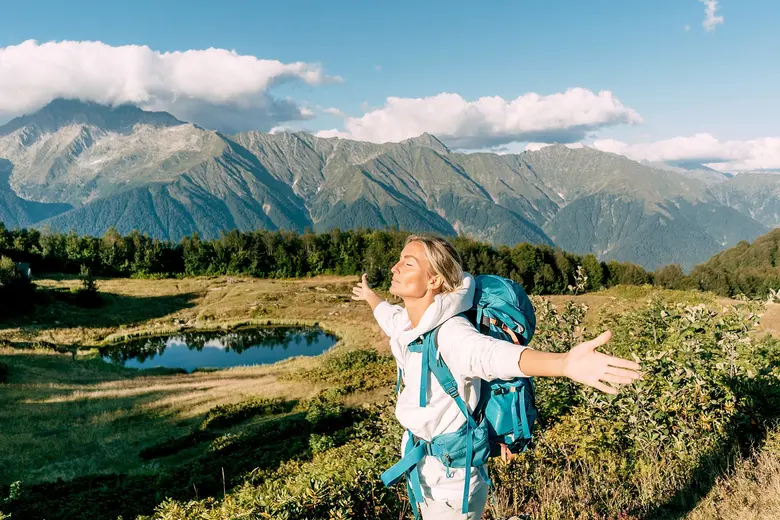 Eine Wanderin mit türkisfarbenem Rucksack steht mit ausgebreiteten Armen und geschlossenen Augen auf einer grünen Bergwiese. Im Hintergrund liegen ein kleiner See und ein imposantes Bergpanorama unter blauem Himmel.