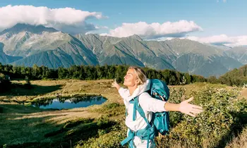 Eine Wanderin mit türkisfarbenem Rucksack steht mit ausgebreiteten Armen und geschlossenen Augen auf einer grünen Bergwiese. Im Hintergrund liegen ein kleiner See und ein imposantes Bergpanorama unter blauem Himmel.