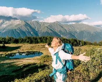 Eine Wanderin mit türkisfarbenem Rucksack steht mit ausgebreiteten Armen und geschlossenen Augen auf einer grünen Bergwiese. Im Hintergrund liegen ein kleiner See und ein imposantes Bergpanorama unter blauem Himmel.