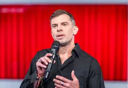 Mann mit kurzem Haar und schwarzem Hemd spricht gestikulierend in ein Mikrofon. Im Hintergrund ist eine leuchtend rote Wand zu sehen.