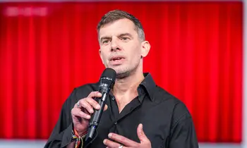 Mann mit kurzem Haar und schwarzem Hemd spricht gestikulierend in ein Mikrofon. Im Hintergrund ist eine leuchtend rote Wand zu sehen.