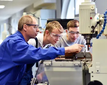 Junge Auszubildende mit älteren Ausbildern an einer CNC-Drehmaschine in einer Werkhalle.