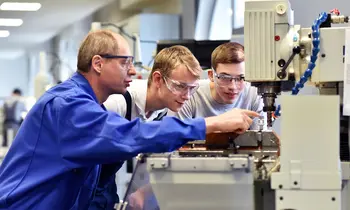 Junge Auszubildende mit älteren Ausbildern an einer CNC-Drehmaschine in einer Werkhalle.