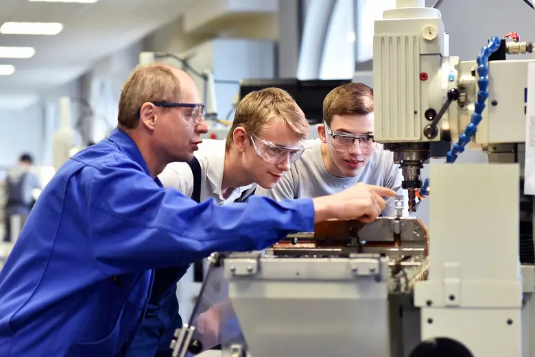 Junge Auszubildende mit älteren Ausbildern an einer CNC-Drehmaschine in einer Werkhalle.