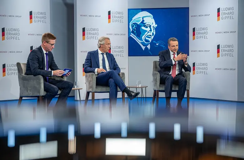 Drei Männer sitzen bei einer Podiumsdiskussion auf einer Bühne mit Ludwig-Erhard-Gipfel-Logos.