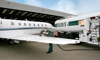 Ein Mitarbeiter in grüner Arbeitskleidung schließt einen Schlauch an den Flügel eines weißen Business-Jets an, im Hintergrund ein Tanklaster mit der Aufschrift Rubis.