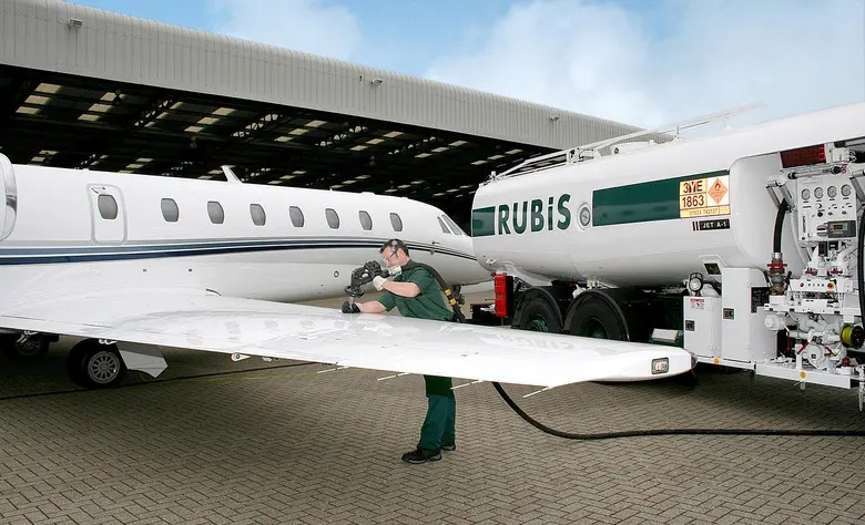 Ein Mitarbeiter in grüner Arbeitskleidung schließt einen Schlauch an den Flügel eines weißen Business-Jets an, im Hintergrund ein Tanklaster mit der Aufschrift Rubis.