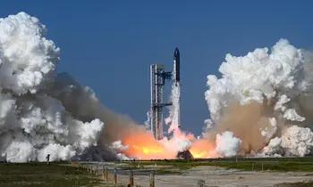 Eine große Rakete hebt vom Startplatz ab, umgeben von massiven weißen Rauchwolken und orangefarbenen Flammen, vor blauem Himmel.