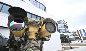 Ein khakifarbenes Sensorsystem mit zwei großen Linsen ist auf einer Halterung montiert, im Hintergrund steht eine Person in Tarnkleidung vor einem modernen verglasten Gebäude mit mehreren gehissten Flaggen.