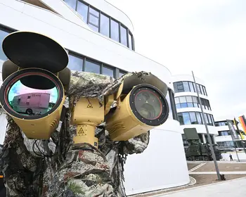 Ein khakifarbenes Sensorsystem mit zwei großen Linsen ist auf einer Halterung montiert, im Hintergrund steht eine Person in Tarnkleidung vor einem modernen verglasten Gebäude mit mehreren gehissten Flaggen.