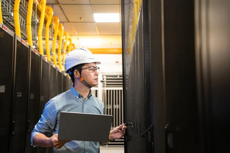 Techniker mit Schutzhelm und Laptop steht zwischen hohen Serverracks in einem Rechenzentrum.