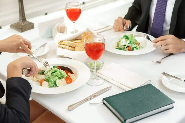 Vogelperspektive auf einen weiß gedeckten Restauranttisch. Links und rechts davon halten zwei Personen in dunklen Businessjacken Besteck in den Händen und essen Salat. Auf dem Tisch stehen zwei Gläser mit rotem Getränk sowie ein Brotkorb. Unten rechts liegt ein grünes Notizbuch.