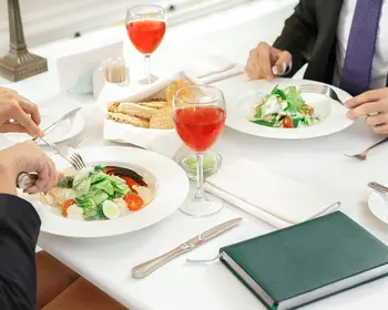 Vogelperspektive auf einen weiß gedeckten Restauranttisch. Links und rechts davon halten zwei Personen in dunklen Businessjacken Besteck in den Händen und essen Salat. Auf dem Tisch stehen zwei Gläser mit rotem Getränk sowie ein Brotkorb. Unten rechts liegt ein grünes Notizbuch.