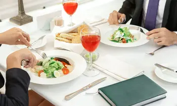 Vogelperspektive auf einen weiß gedeckten Restauranttisch. Links und rechts davon halten zwei Personen in dunklen Businessjacken Besteck in den Händen und essen Salat. Auf dem Tisch stehen zwei Gläser mit rotem Getränk sowie ein Brotkorb. Unten rechts liegt ein grünes Notizbuch.